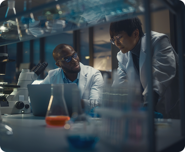 Two scientists in lab coats reviewing research together beside a microscope and laboratory equipment.